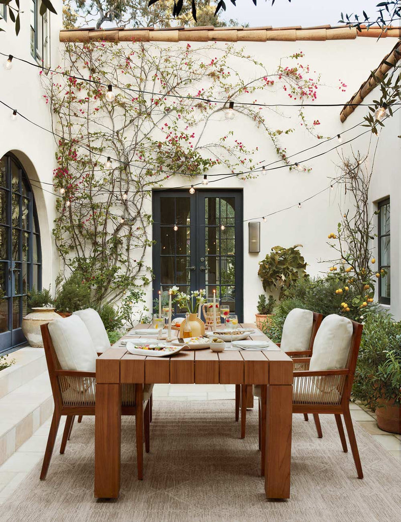 A teak wood dining table has four wooden arm chairs with white cushions anchor an outdoor courtyard patio. Hanging lights are over the table and herbs and vegetables line the courtyard.