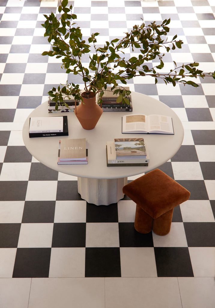A black and white checkerboard tile floor grounds an entryway with a round off-white table with books displayed on top and a brown velvet footstool.