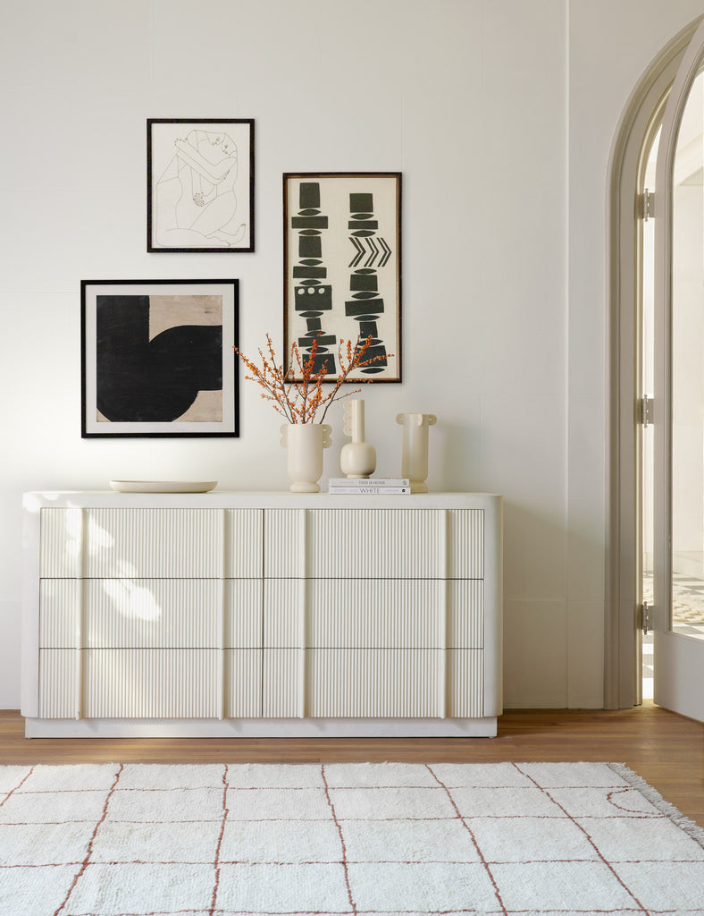 A white curved dresser with reeded front drawers adds storage in an entryway. On top of the dresser sits a white low bowl and three white ceramic vases of varying heights. Above the dresser hangs three modern black and white paintings.