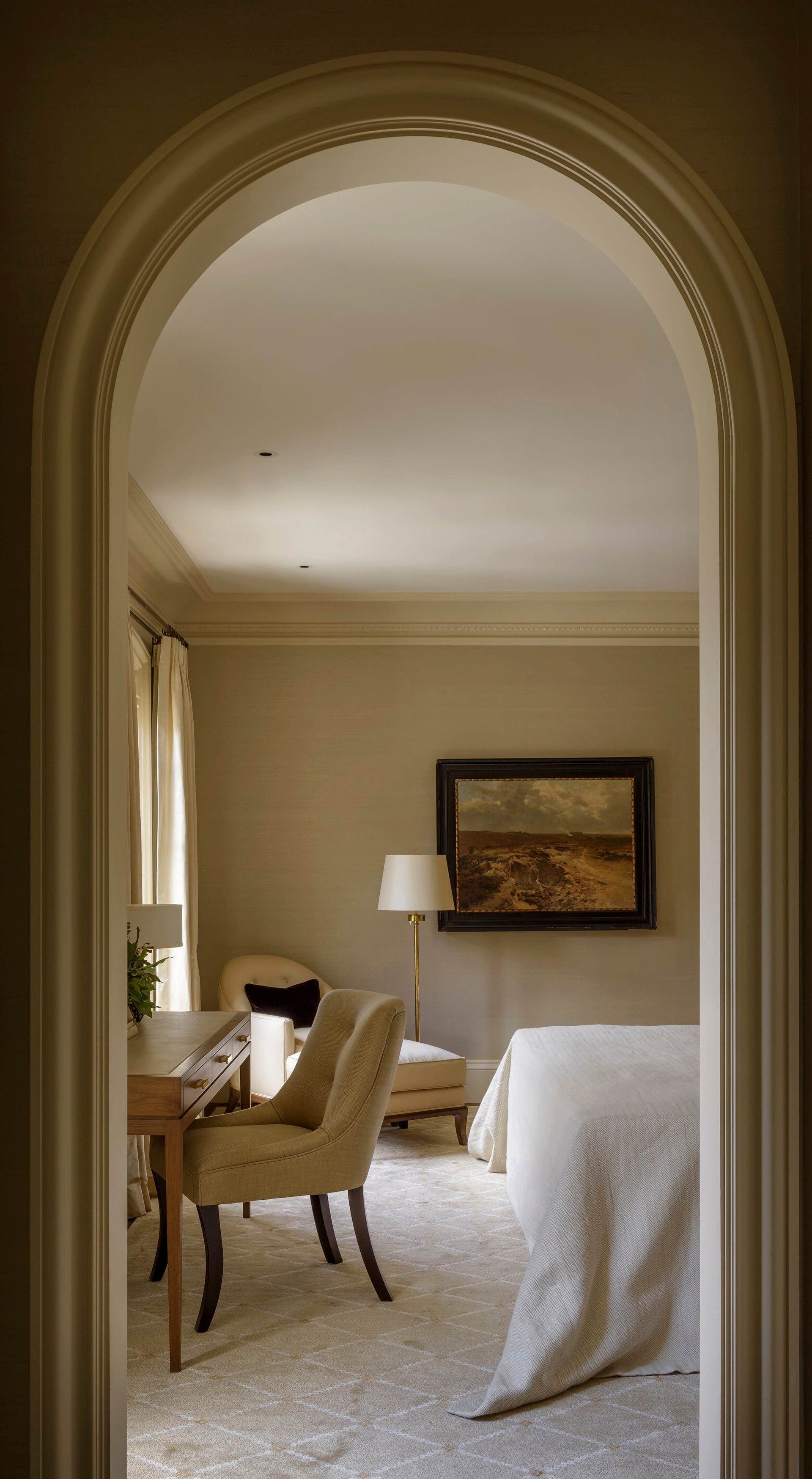 A bedroom through an archway has a bed with white bedspread, a light wood writing desk and gold upholstered chair and a rounded ivory chair in the corner with an ottoman.