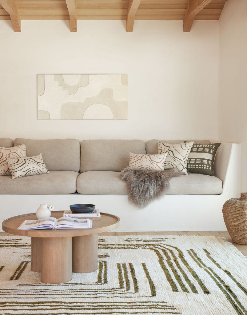 A white built-in sofa has light tan cushions and neutral throw pillows with a wavy design and a fuzzy blanket. A small round light wood coffee table sits in front of hte sofa on a neutral rug with a dark green geometric line design.