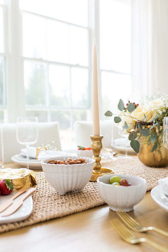 Nuts and fruit sit in textured white bowls on a light wood dining table. Behind them sit a tall white taper candle in a gold holder on a woven seagrass runner.