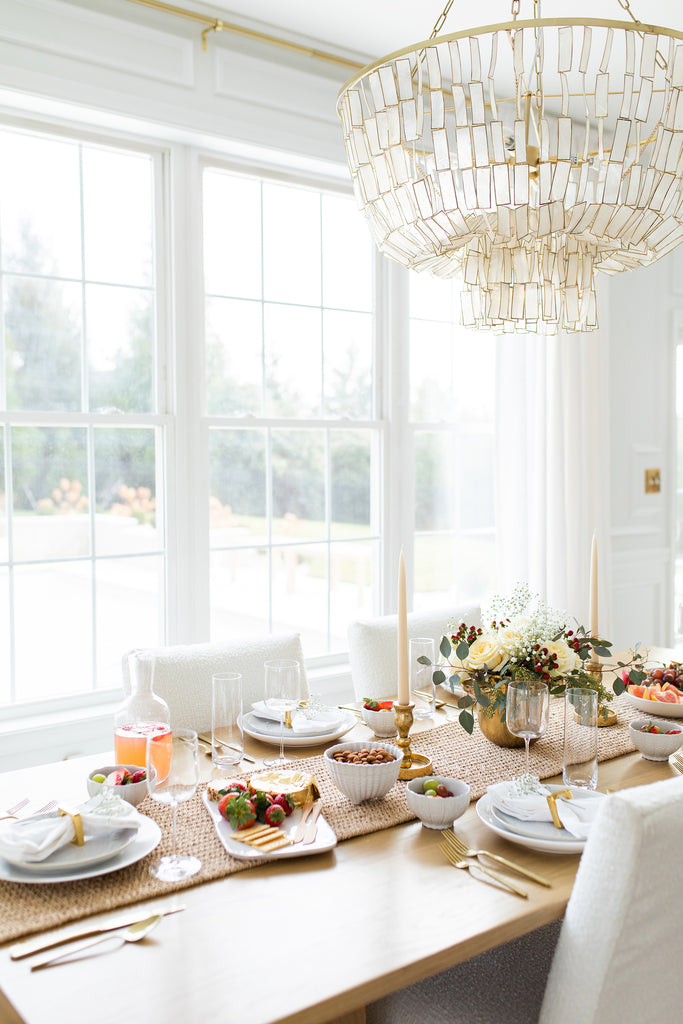 A full view of Nabela Noor's dinner table with white slipcovered dining chairs around a light wood rectangular dining table. On top sits white place settings with gold flatware and bowls of nuts and fruit.