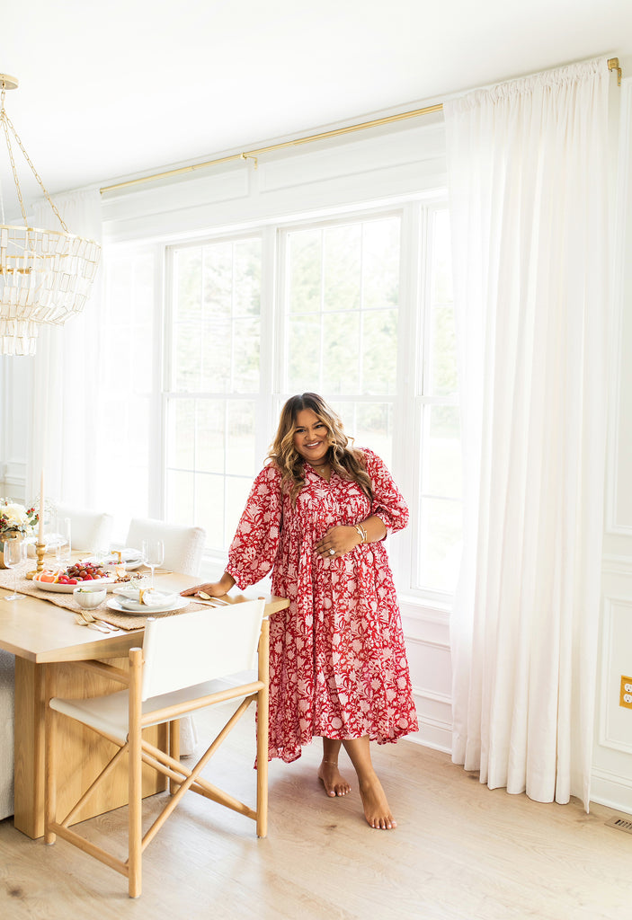 Creator Nabela Noor in a red midi floral dress stands leans on her light wood dining table surrounded by slipcovered white chairs and a wood framed arm chair at the head of the table.