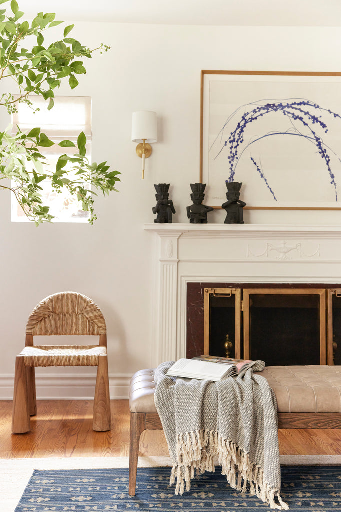 A chunky light wood and rattan woven Heidy accent chair sits next to a leather upholstered bench with wooden legs with a blanket draped over it. The chair sits next to a white fireplace with a large blue and white painting over it and three black statues on it.