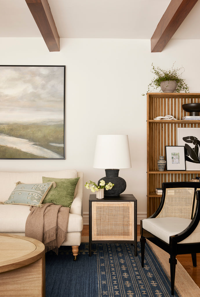 An ivory linen sofa sits next to a black square side table with a cane door. A black lamp sits atop the table. A black and cane accent chair sits next to the table and a curved bookcase is behind it.