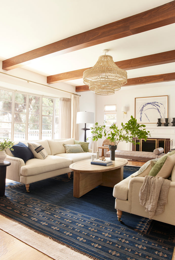 Two ivory linen Fabienne sofas sit across from each other atop a navy wool area rug. A light wood oval coffee table sits in between and a beaded rattan chandelier by Arteriors hangs overhead.