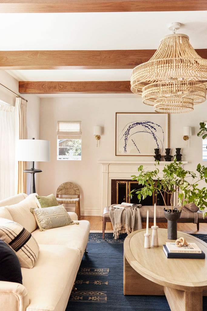 A bead and rattan Arteriors Tulane chandelier hangs above a living room with an ivory sofa, navy blue area rug, light wood oval coffee table, and light brown leather upholstered bench.