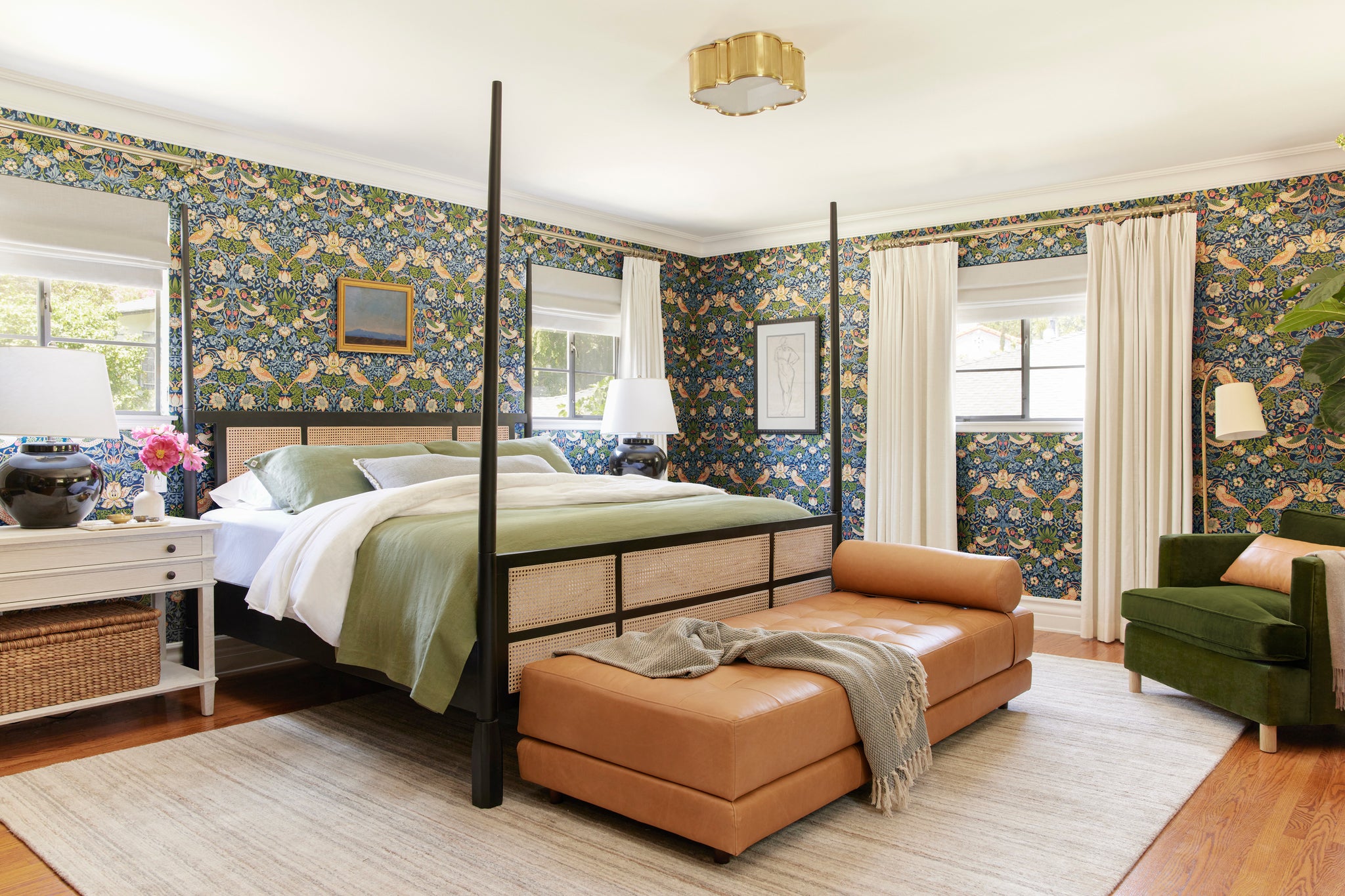 Morris & Co. Strawberry Thief wallpaper hangs on the walls of this bedroom. A black and cane Kaira 4-poster bed is in the middle with sage green bedding. A camel leather chaise lounge sits at the end of the bed. Across from the bed is a dark green velvet armchair.