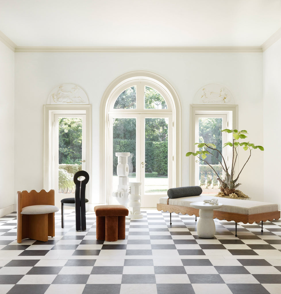 The Ripple stool, black Whit dining chair, brown velvet Clover stool, white Toivo sculptural pedestals, and the Rise wood and linen daybed from the Sarah Sherman Samuel and Lulu and Georgia collection are displayed in a room with a black and white checkered floor.