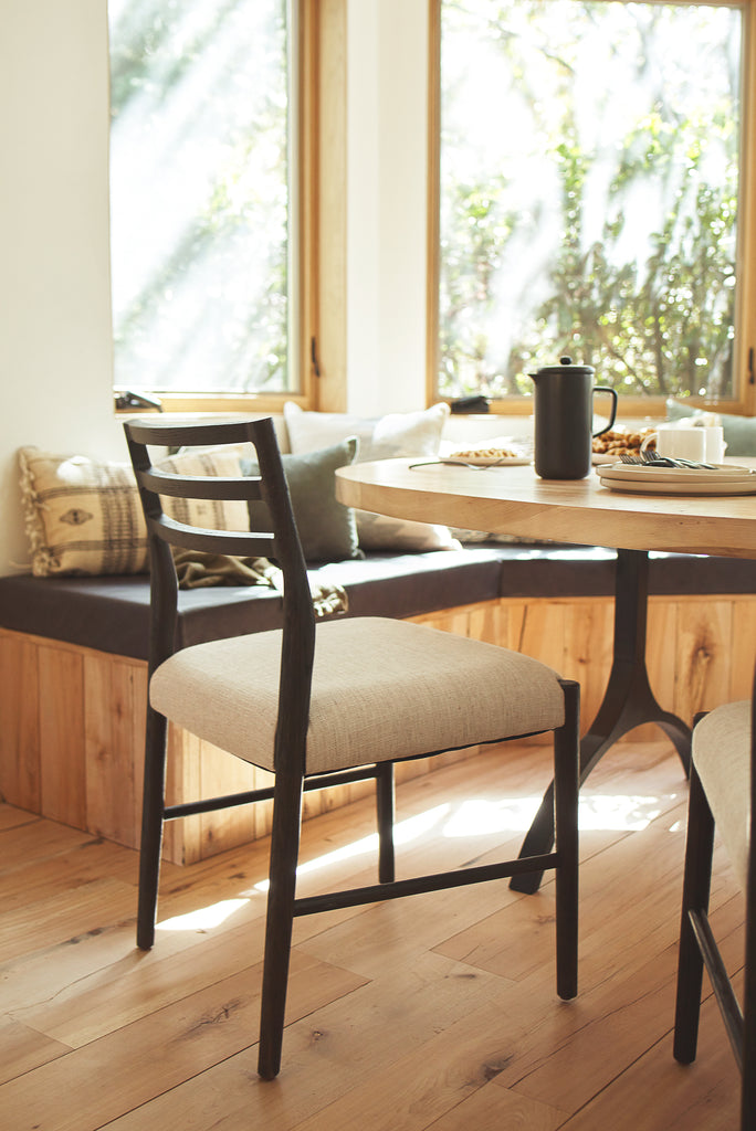 A modern black wooden dining chair with open back slats sits at a round light wood dining table with a black iron base. Across from the chairs is a built-in banquet covered in light wood paneling and topped with a thick gray cushion and neutral throw pillows.