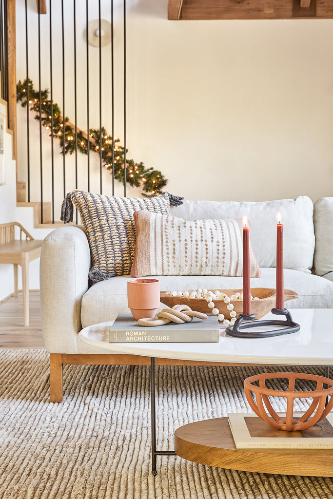 A black SIN Duo Candlestick Holder has two lit red tapered candles on an oval marble, iron and wood coffee table in front of an ivory linen sofa in this holiday living room.