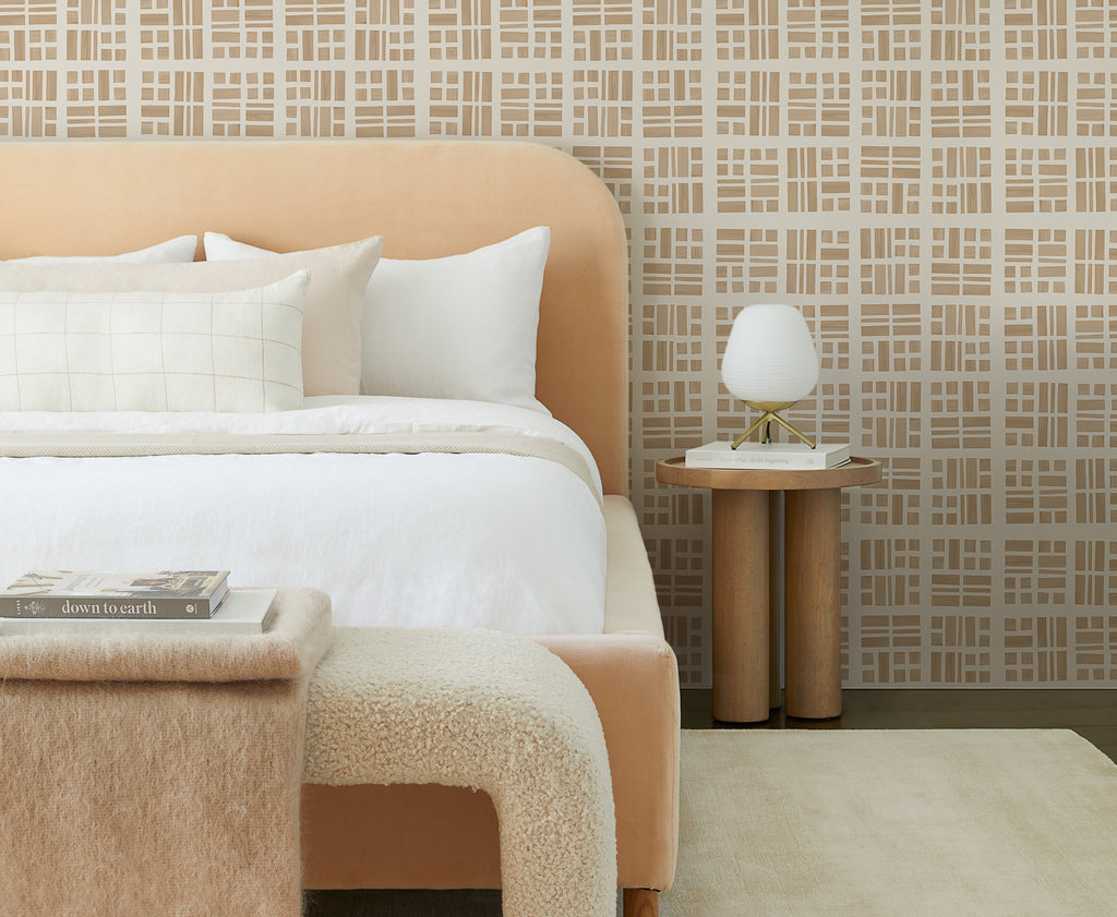 A simple yet plush creamy area rug sits underneath a blush velvet upholstered platform bed with cream colored bedding. At the foot of the bed is a rounded boucle bench. A modern grid-patterned wallpaper in tan and white hangs as a backdrop.