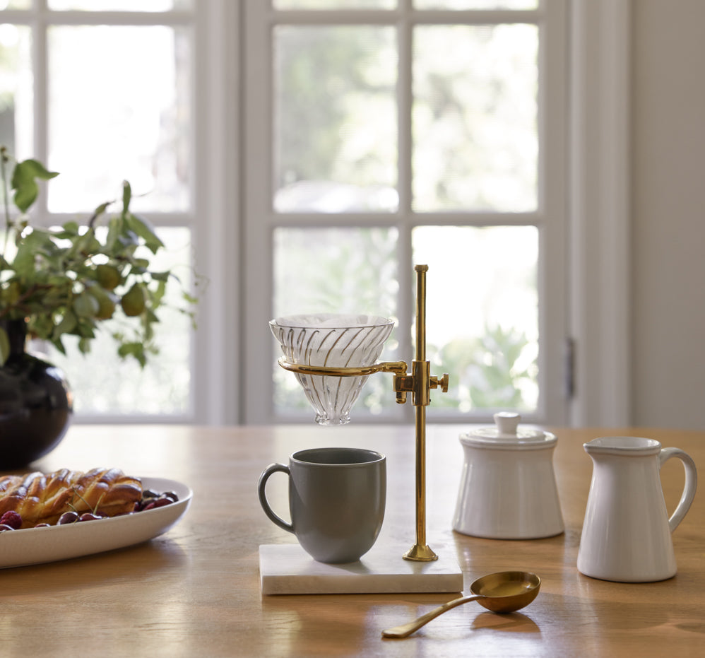 A brass and marble pour-over coffee stand has a dark green mug underneath it and a gold spoon.