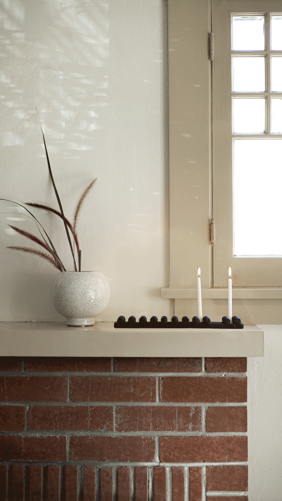 A modern, low black Menorah holds white lit tapered candles atop a red brick fireplace with neutral stone mantel.
