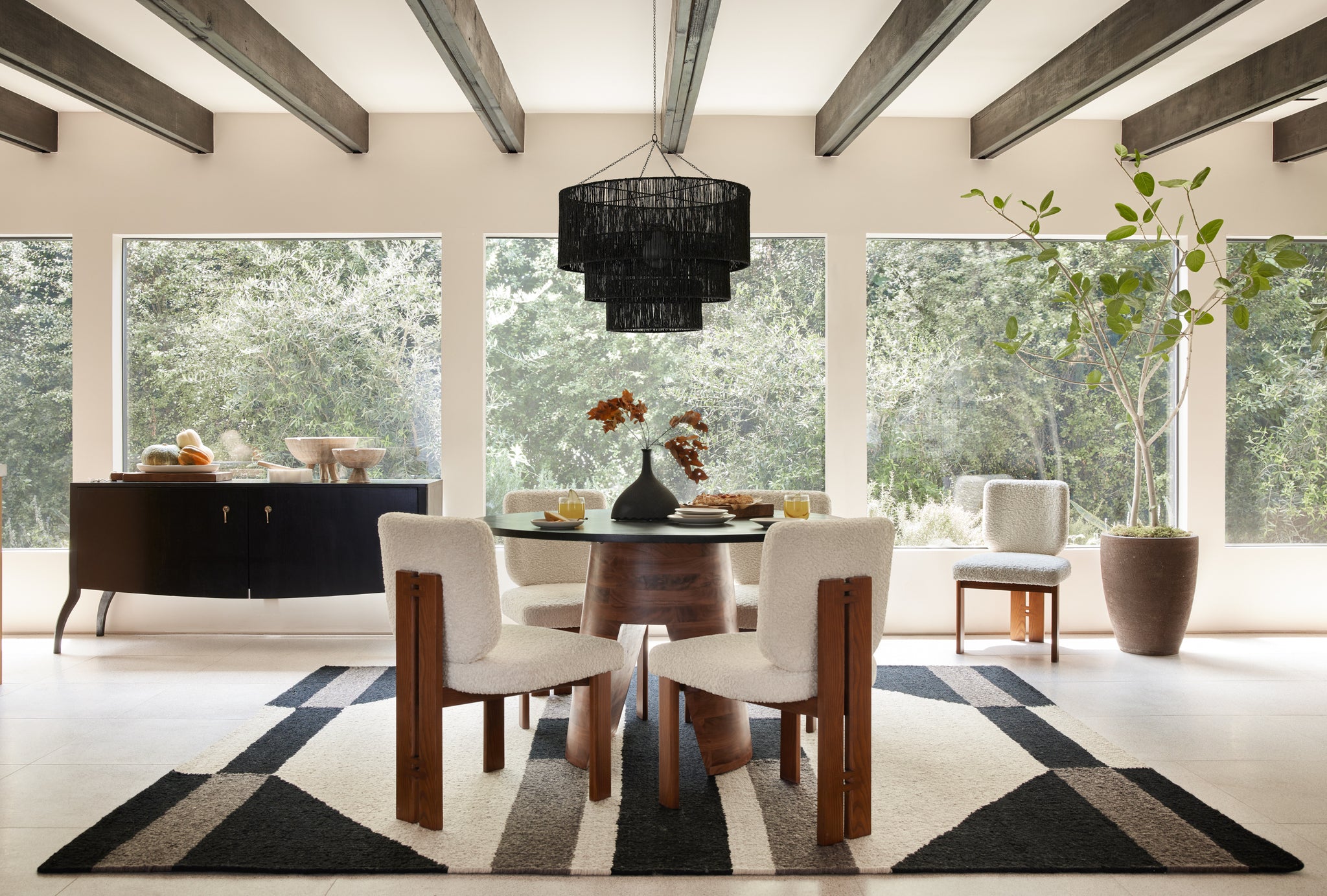 The black woven Sayan tiered chandelier pendant light hangs above a round dining table with a black top and wooden pedestal base. Four white boucle dining chairs with wooden backs and legs surround the table while a fifth chair is off to the side next to a potted tree. A black sideboard sits against a wall of windows.