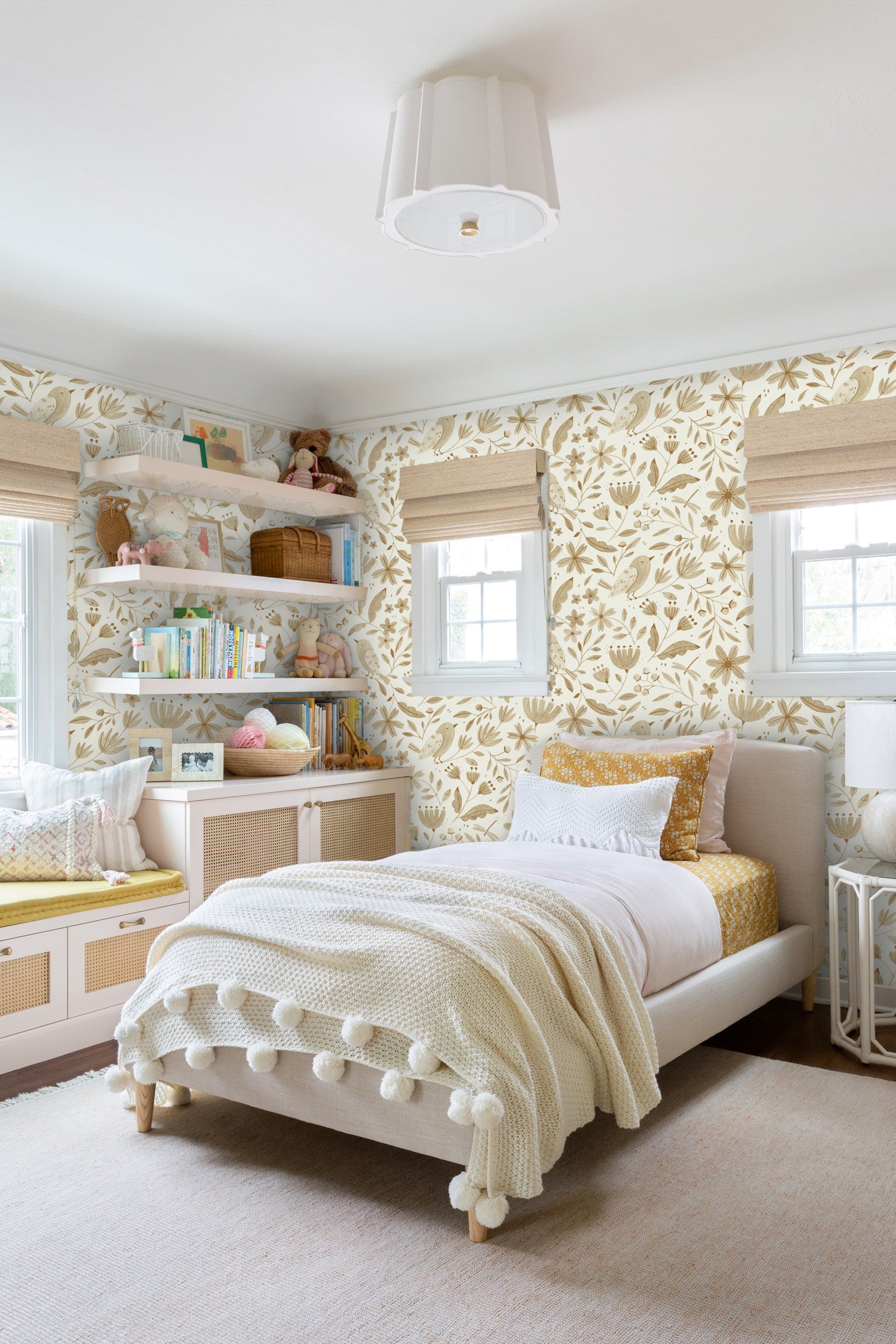 The neutral botanical Garden Birds wallpaper from Rylee + Cru anchors this kids' room with a twin bed covered in white and gold bedding and built-in cabinets with cane fronts and open shelving holding toys and books.