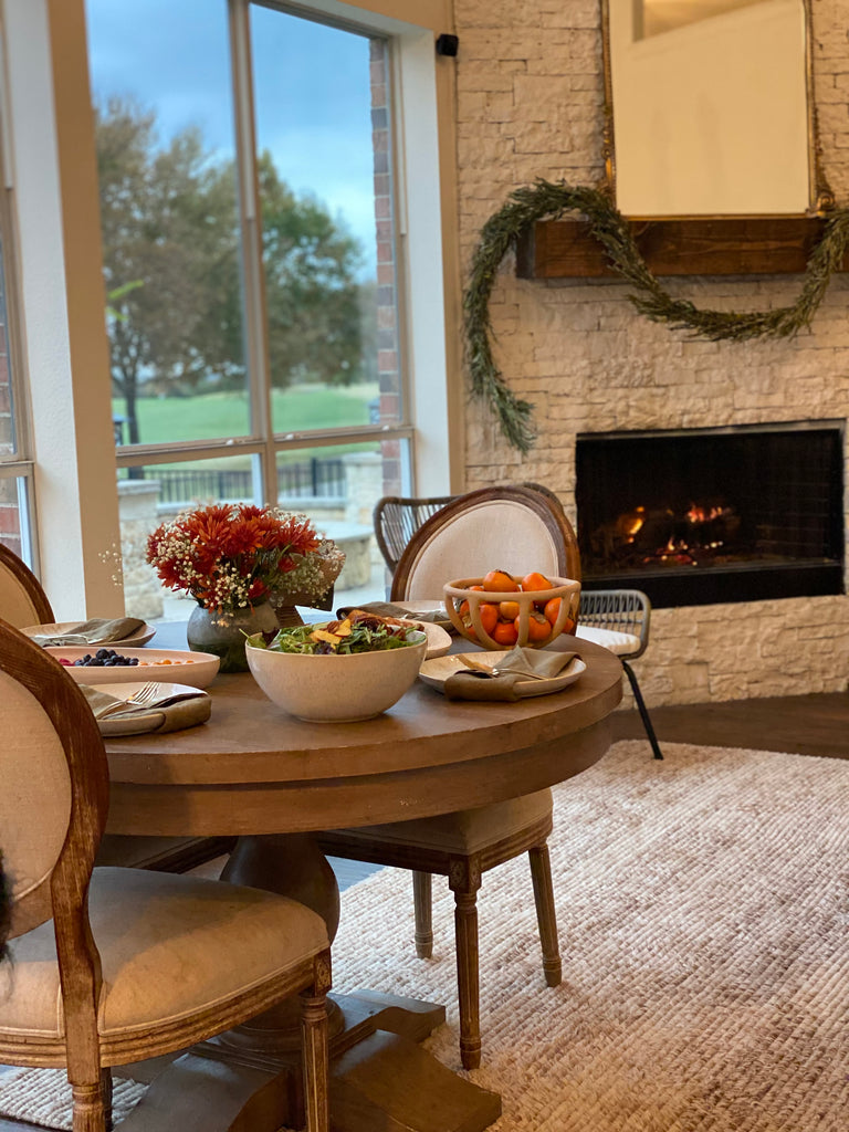 Ascia Sahar's holiday spread is set on a round wooden pedestal table with rounded upholstered dining chairs with wooden frames in front of a stacked stone fireplace.