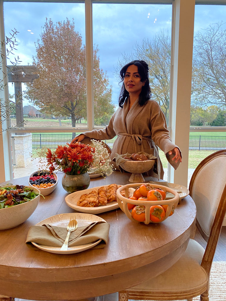Influencer and blogger Ascia Sahar stands over her holiday table complete with a basket of persimmons, handpies, a winter salad, fresh fruit, a bread basket and an orange and white floral arrangement.