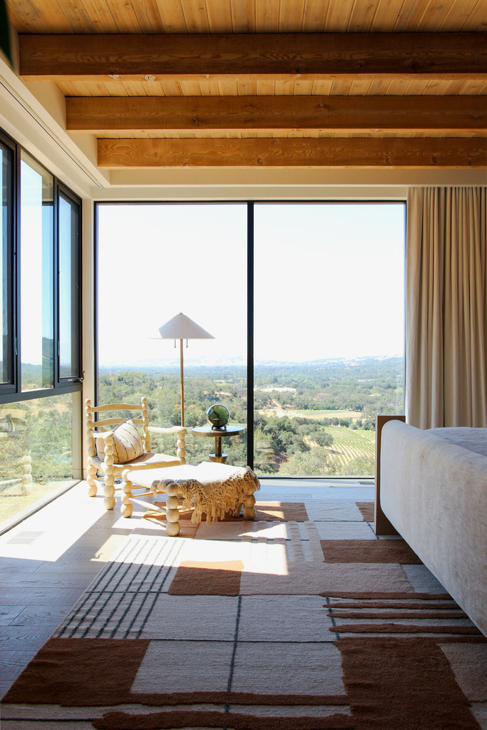 A light-filled bedrooms with huge windows in the corners overlooking a valley have a carved wooden chair with matching ottoman atop a brown and neutral grid rug with an upholstered footboard.