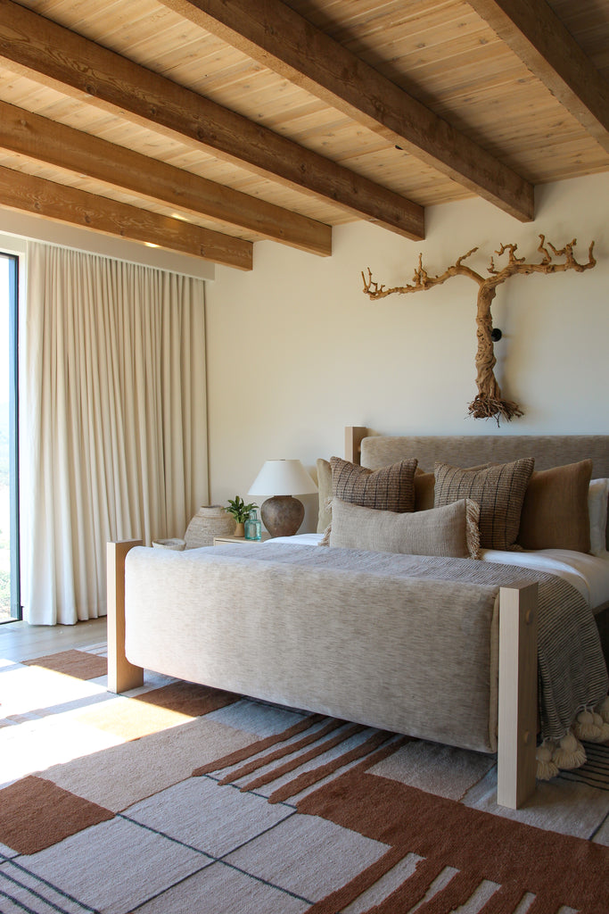 A light tan upholstered bedframe has neutral colored bedding and sits atop a brown and neutral grid rug. A tree branch hangs on the wall above the bed.