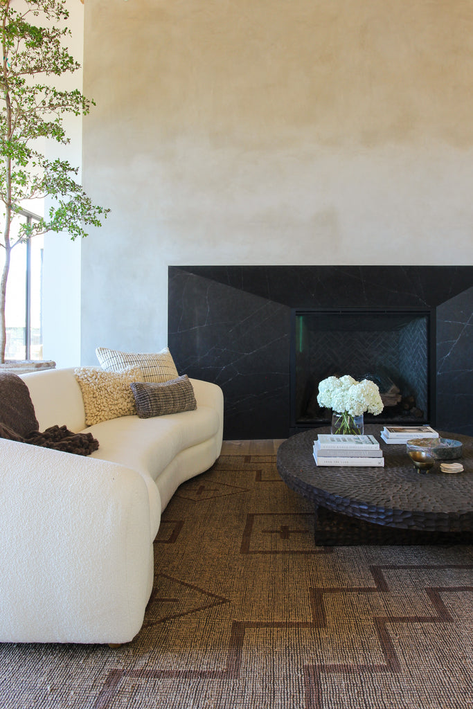 A curved white boucle sofa sits opposite a round hammered metal coffee table in front of a black stone fireplace.