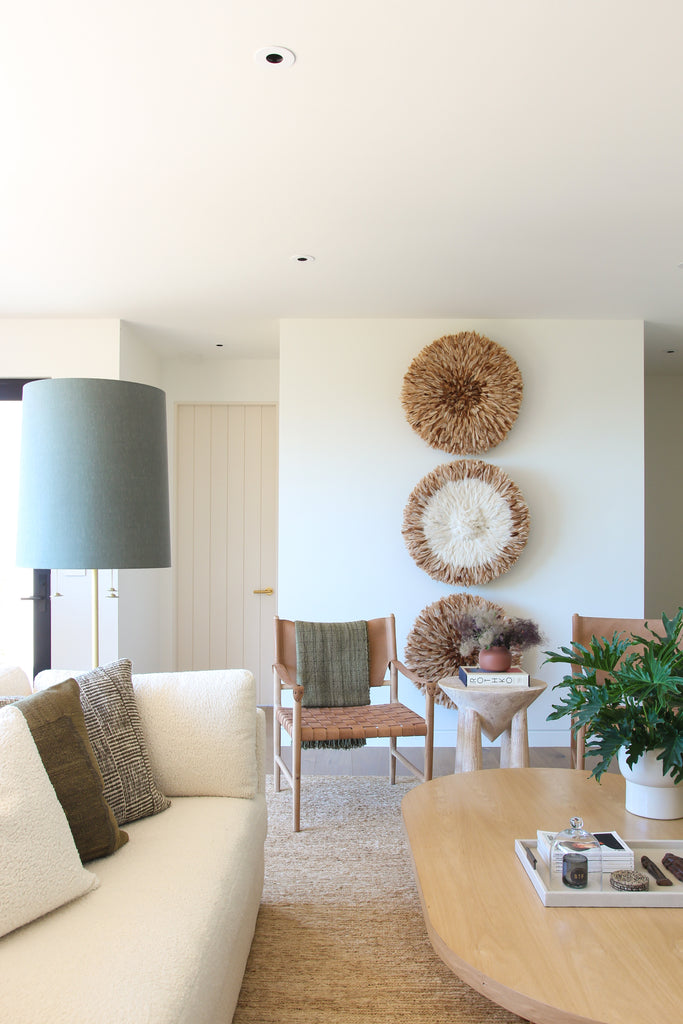 A bright living room has an ivory sofa with a light wood coffee table and two woven leather arm chairs with three round woven wall hangings behind the chairs.