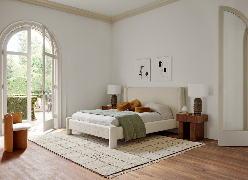 The white boucle Hyvaa bed has neutral bed linens and a green duvet draped across with three copper velvet round throw pillows stacked on it. Two wooden Merritt nightstands flank the bed with wooden table lamps. The Ripple chair sits across from the bed. All furniture was designed by Sarah Sherman Samuel for Lulu and Georgia.