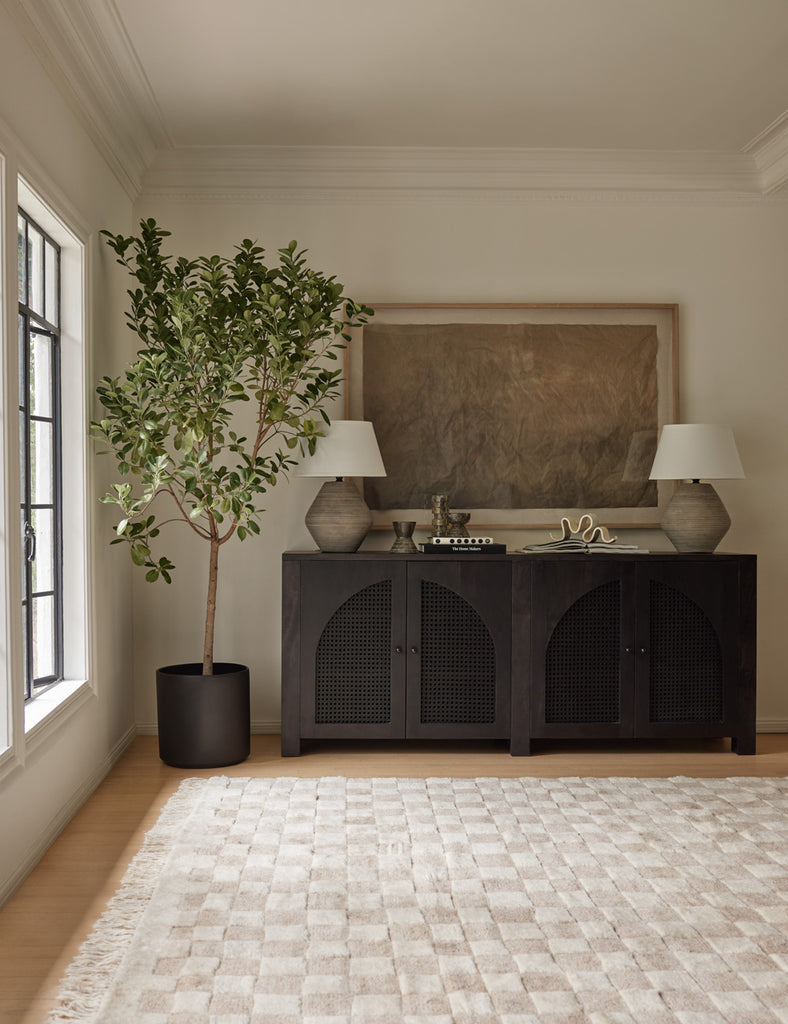 The tan and ivory Irregular Checkerboard Rug by Sarah Sherman Samuel sits in front of a dark wood sideboard with arched cane paneling and a potted tree.