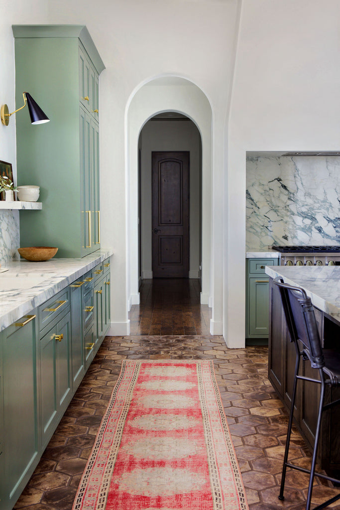 Green cabinets with marble countertops and backsplash and gold hardware define Jamie Lynn Sigler's kitchen. A red medallion runner rug lays between the cabinets and island on a brown hexagon tiled floor.