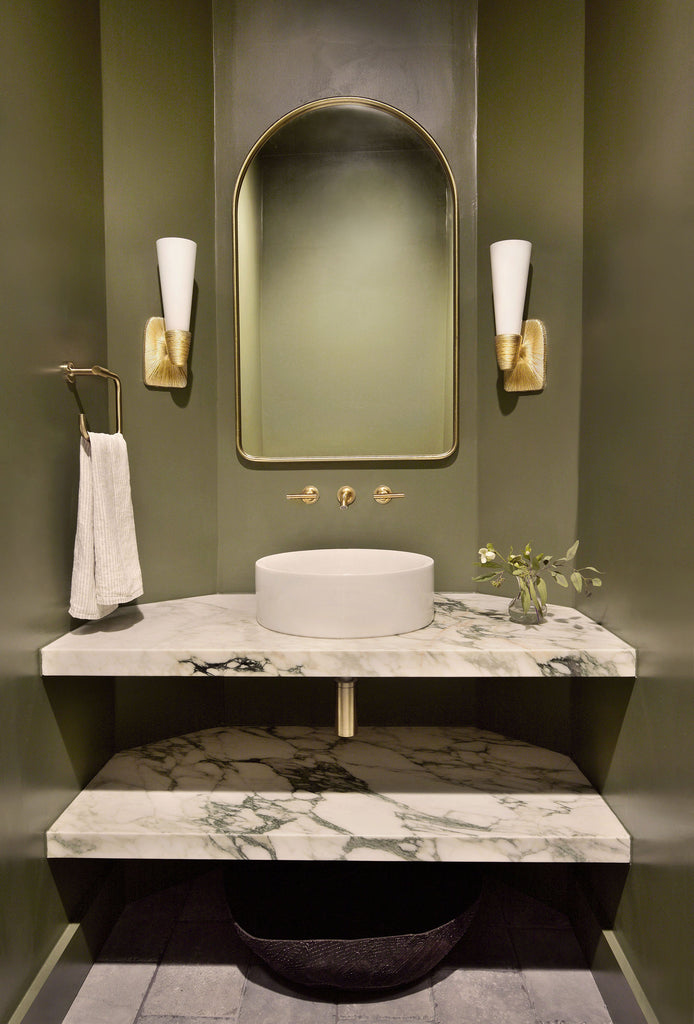 A marble bathroom vanity with a white round vessel sink and gold hardware stand out against the moss green walls in this bathroom. A gold framed mirror and two gold sconces hang above the vanity.