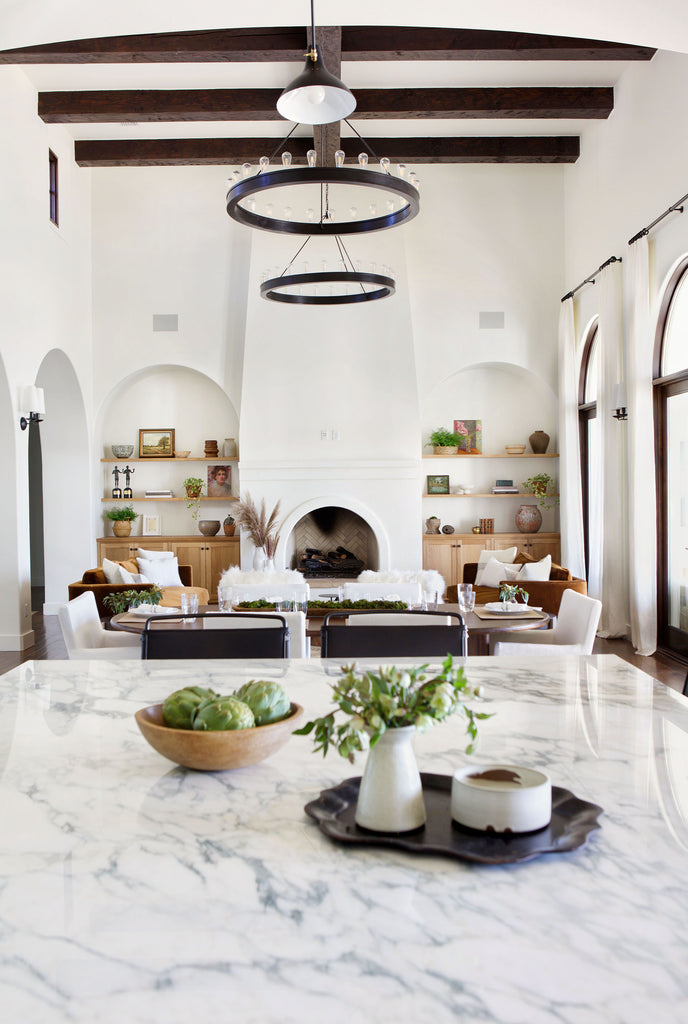 A marble countertop with a bowl of artichokes and tray with a small vase and coasters overlooks the dining and living room of Jamie Lynn Sigler's home. Two large round chandeliers hang over the space and a large fireplace flanked with two built-in arches house light wood shelves and storage cabinets. 