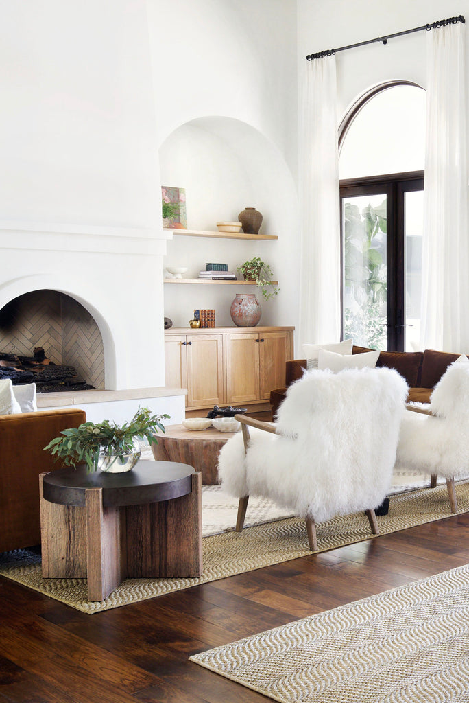 Two white fuzzy Kora accent chairs with wooden arms and legs flank two copper Belmont velvet sofas. A large arched fireplace sits across from the chairs and a round chunky wooden end table next to one of the chairs has a vase holding branches in water.