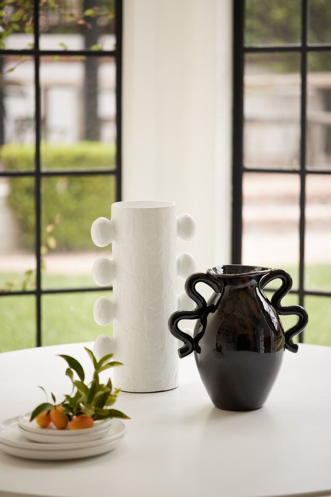 A rounded modern matte white vase sits next to a shiny black vase with wavy handles. A stack of small plates with persimmons sits next to the vases.
