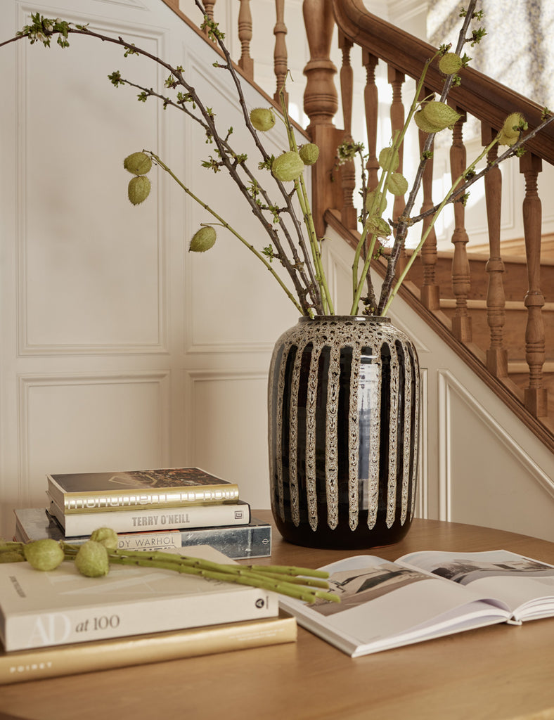 A tall ceramic black vase with a neutral drip glaze holds tall branches and stems and sits atop a wooden table with books.