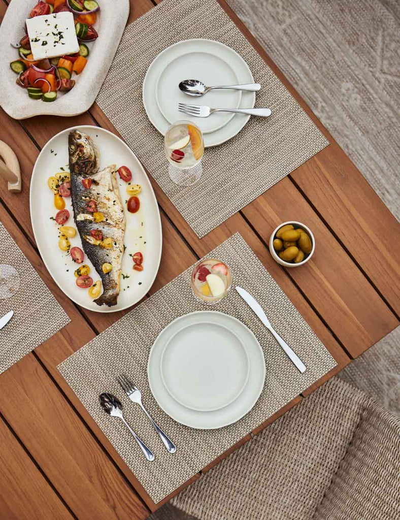 A teak dining table holds three place settings with tan woven placemats, white plates and silver flatware. A cooked fish with tomatoes is served on a white oval serving platter and vegetables and a block of feta cheese are served in another platter.