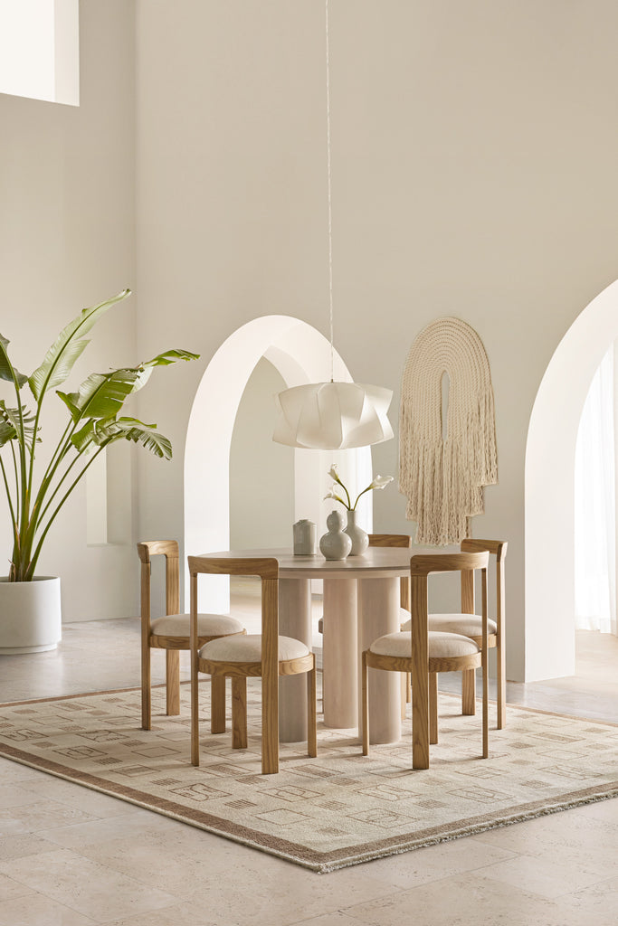 Curved wooden modern dining chairs surround a light wood pedestal round dining table in a light and airy dining room. A neutral patterned rug grounds the space and a modern white sculptural pendant light hangs above the table.