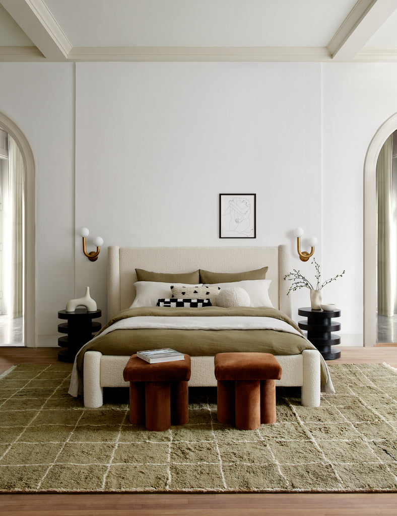 The white boucle Hyvaa bed is covered in moss green bed linens and black and white throw pillows. On either side of the bed are the black Pentwater round side tables and at the foot of the bed are two brown velvet Clover stools. All furniture was designed by Sarah Sherman Samuel for Lulu and Georgia.