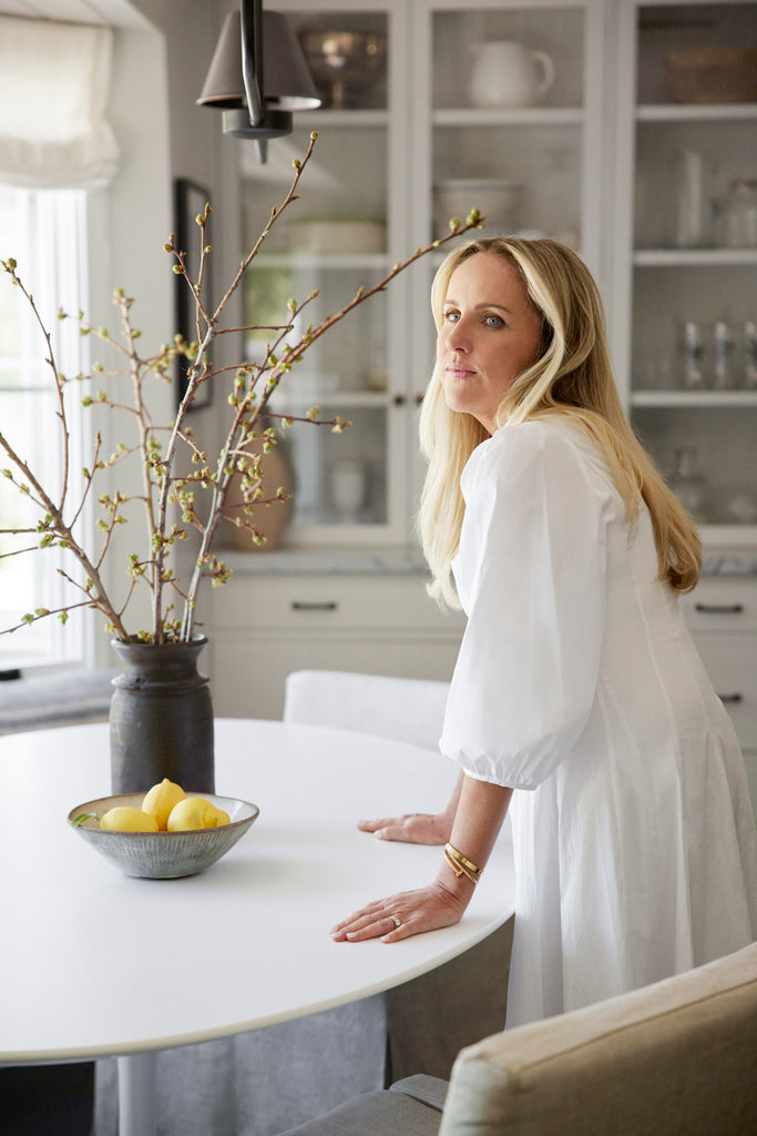 Designer Sarah Solis leans against the round white dining table in her dining nook. On the table is a milk jug-shaped black vase holding tall stems with green buds and a low blue and white ceramic bowl holding lemons.