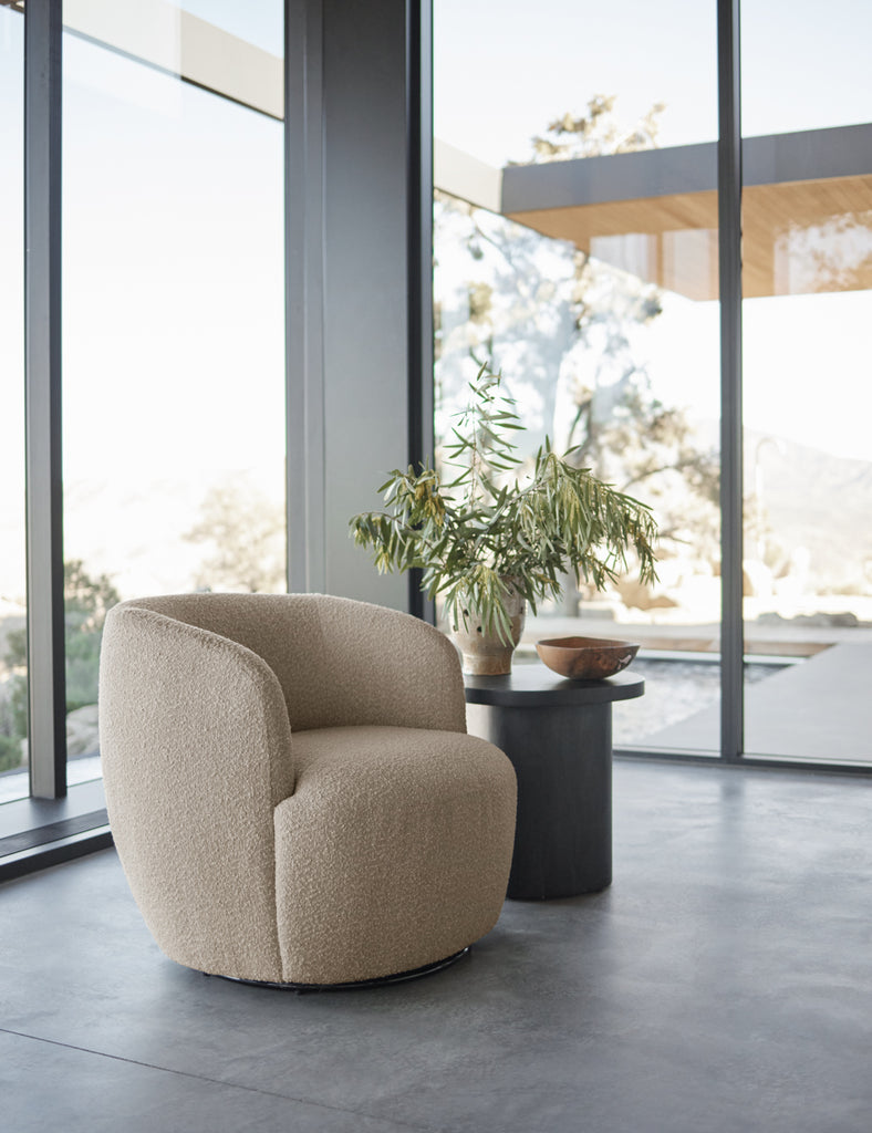 A curved taupe boucle arm chair sits next to a small round dark wood side table. A Namu Home Goods wooden bowl sits on the table.