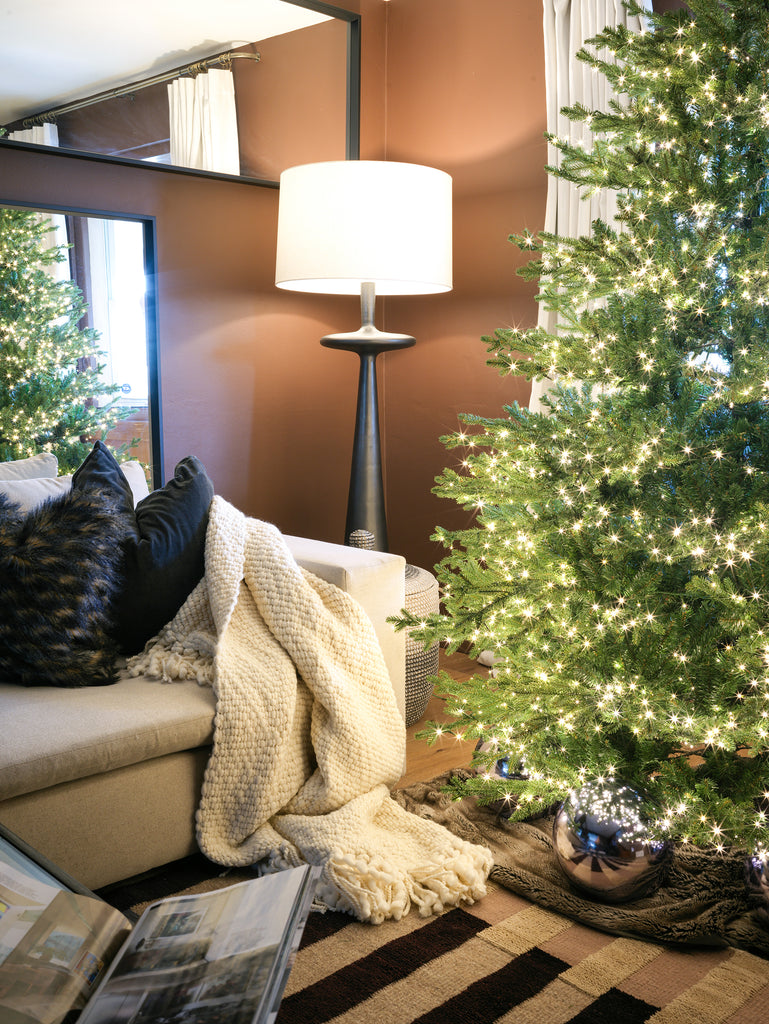 A woven cream throw blanket is draped over a light gray sofa next to black throw pillows. The sofa is next to a lit Christmas tree. A tall black lamp with a white shade stands in the background.