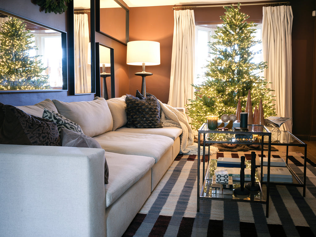 A modern light gray sofa sits across from a black metal and glass tiered coffee table. The neutral striped Otti rug grounds the space and a lit Christmas tree twinkles in the background.