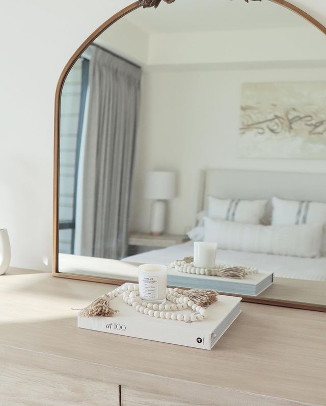 An arched gold framed mirror leans against a wall on top of a light wood dresser. A book, beads and candle sit on the dresser. The mirror reflects the neutral upholstered bed and bedding.