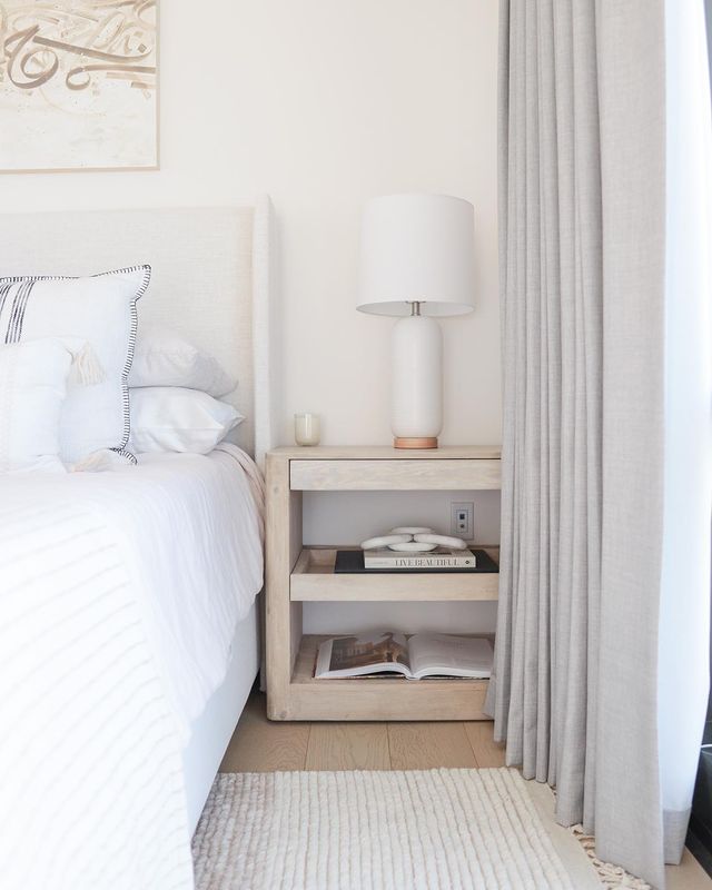 A square light wood nightstand sits next to an ivory upholstered bed with ivory bedding. On top of the nightstand sits a tall cylindrical ivory lamp and two books sit on the nightstand's two shelves.