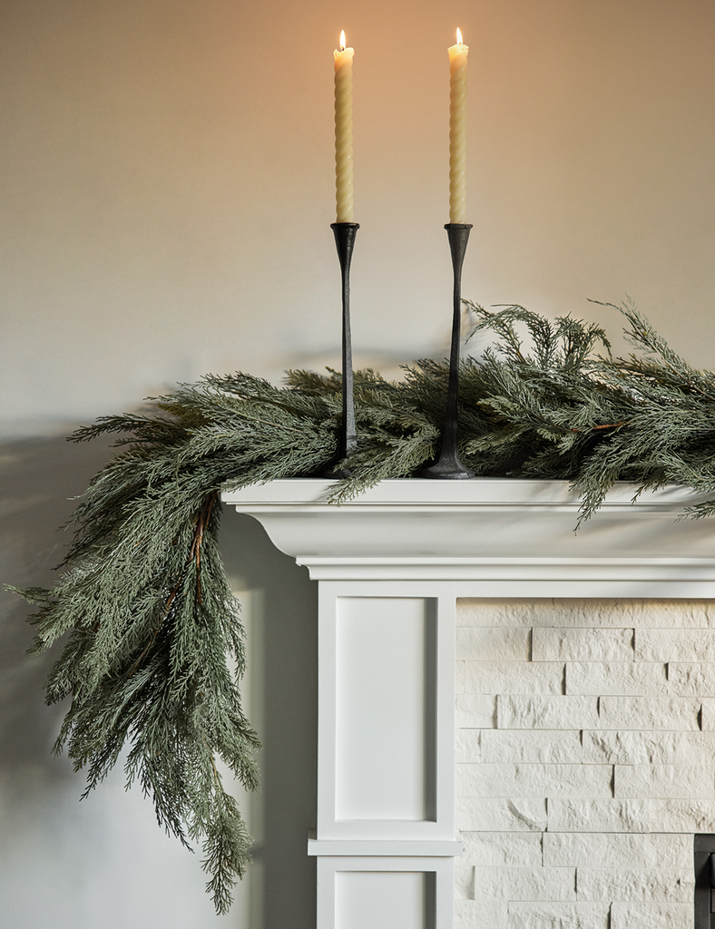 Two lit taper candles in black candle sticks on a mantel with fresh garland.