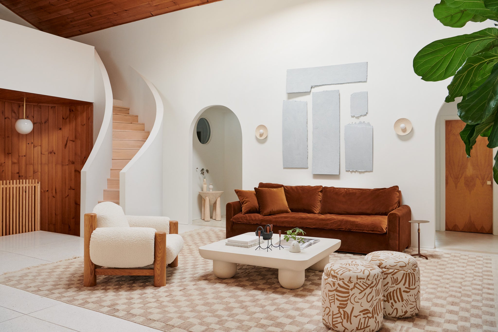 A rust colored modern sofa sits in an open living room with a white square coffee table, sherpa accent chair, and two round rust and white patterned ottomans.