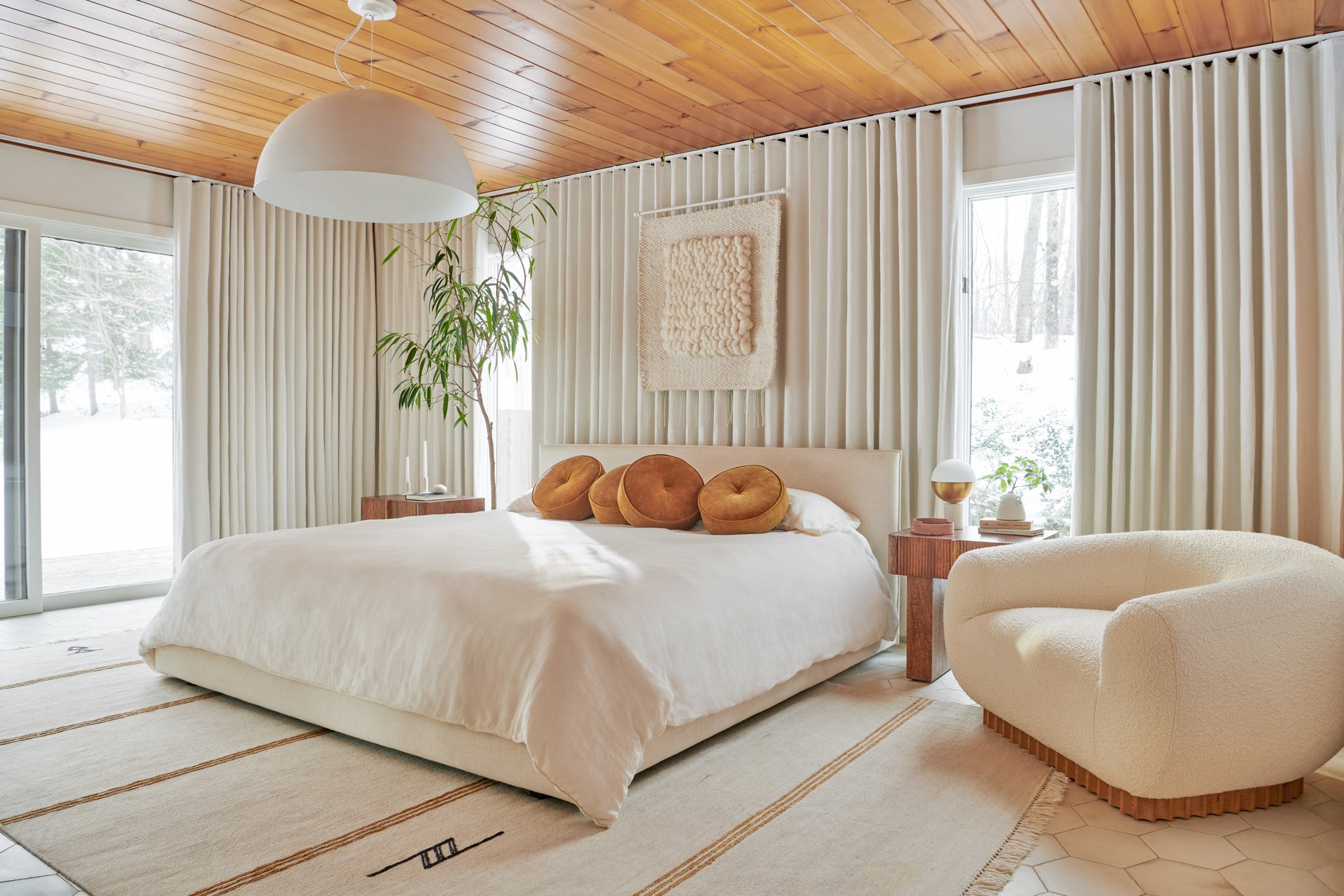 A light bedroom has a low upholstered headboard, cream colored bedding with a pile of round rust colored pillows and walls covered in ivory curtains. A rounded boucle chair sits next to the bed.