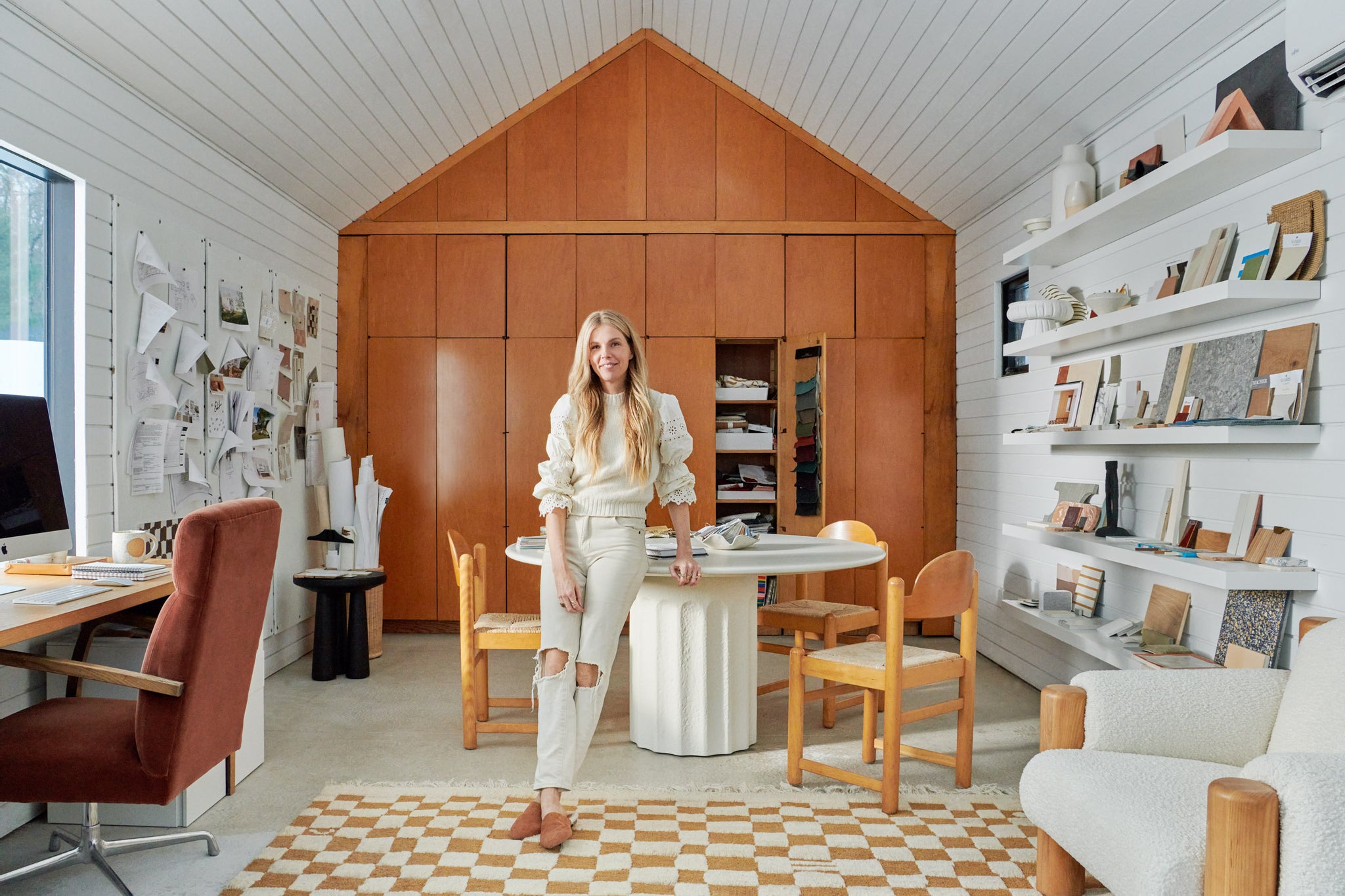 Interior designer Sarah Sherman Samuel stands in her design studio leaning against a round white table surrounded by wooden chairs with woven seats. She is surrounded by swatches and samples on the walls and table.