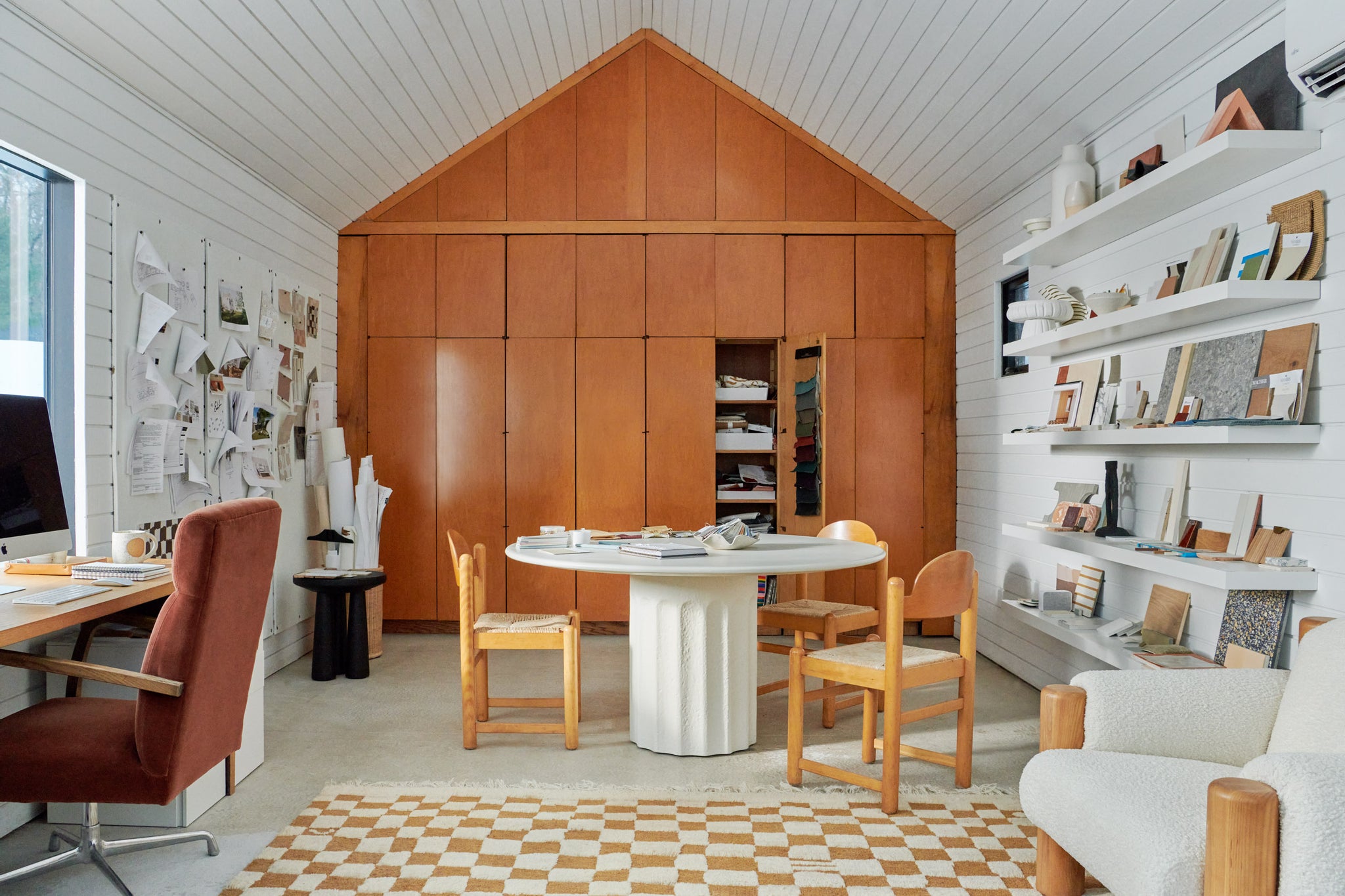 The ochre and white Irregular Checkerboard Rug by Sarah Sherman Samuel is displayed in a design office with a round white table surrounded by three wooden chairs and design samples laying about.
