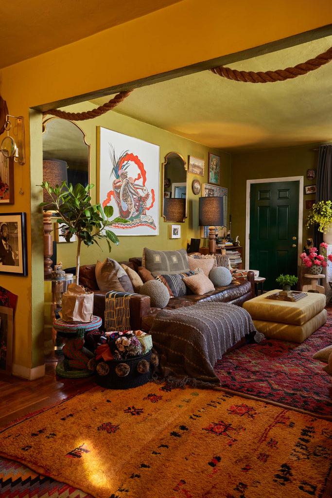 Florist and artist Maurice Harris's living room has a dark brown leather sofa covered in throw pillows, a gold upholstered ottoman, and red Moroccan rugs layered on top of each other.